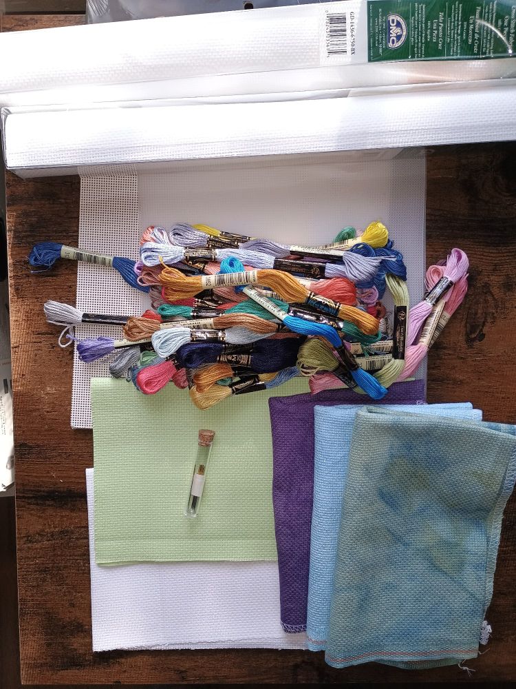 Picture of cross stitch supplies. Loose Aida fabric in lime green, mottled purple, light aqua with opalescent shimmers, blue green, white and 2 packaged rolls of white. A heap of many different colored skeins of floss. Perforated paper and plastic canvas. A tube of fancy Tulip brand needles. 