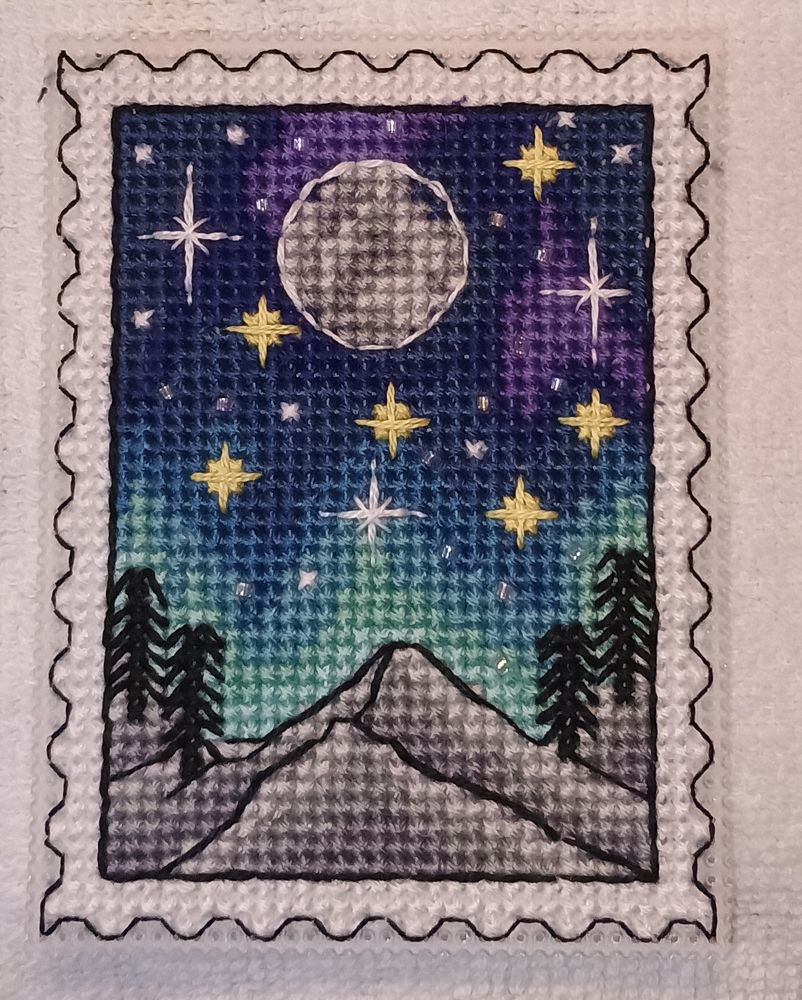 Northern Lights cross-stitch scene. Stitched on 14 count plastic canvas, approximately 2 inches by 3 inches. Piece has scalloped white border. Sky is in shades of purple and dark blue at top, gradually changing to lighter shades of blue and blue-green as you go down. A full moon in shades of gray is in the top middle. Gray and white mountains are at the bottom of the scene. There are two trees on either side at the bottom. In the sky there are white stars, yellow stars and both white dots and random tiny crystal beads representing stars. 