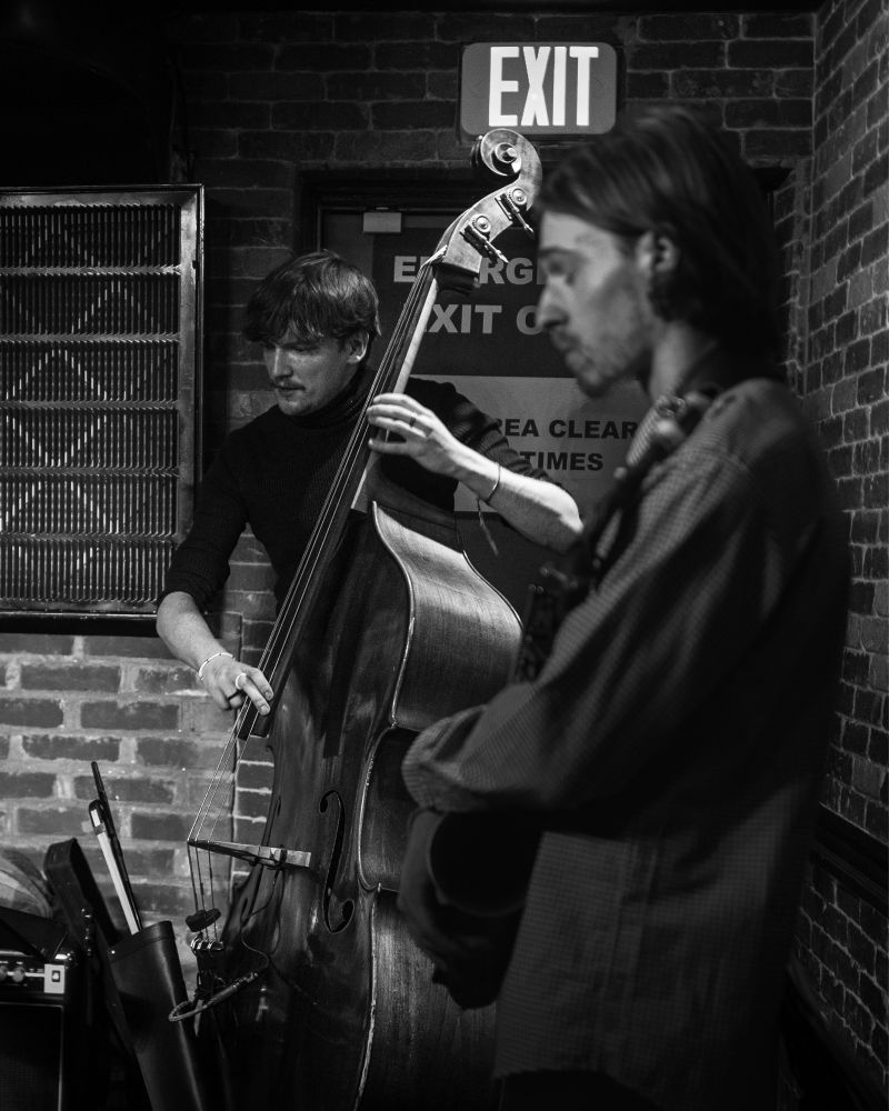 In the foreground there is a blurry person intently listening to the main focus of the image, the upright bass. The bass player is a tall, young man with light skin. He is in front of a rear exit door that has a very bright ‘Exit’ sign above him.