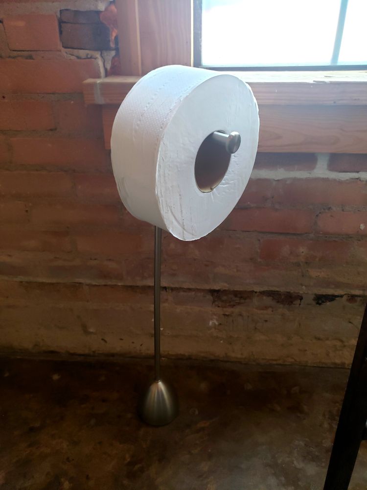 A freaking huge roll of toilet paper dwarfing the toilet paper stand it's on up against the wall near a window