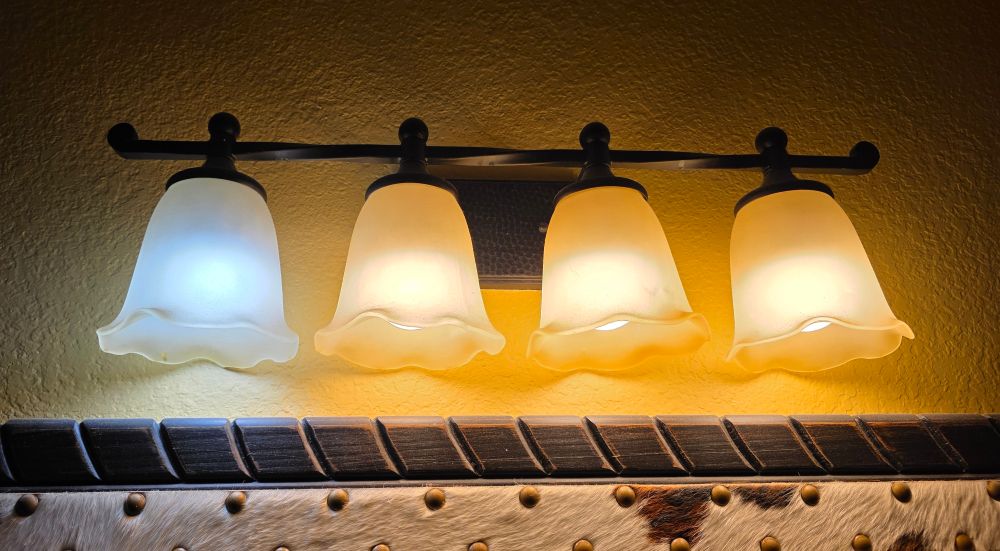 A fixture in a hotel bathroom, three of the bulbs are very warm color temperature, one is very cool. There is a cowhide and brass tack trimmed wooden mirror frame below. Intriguing.