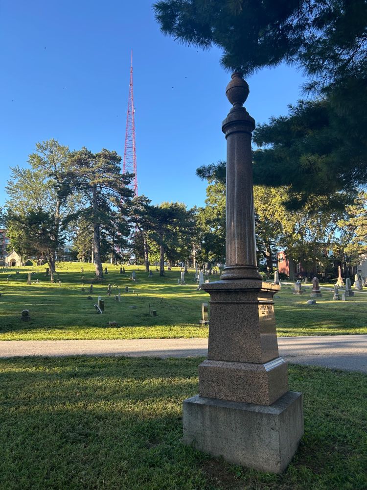 Red granite  column shaped monument in an old cemetery with a red broadcast tower standing tall against a blue sky.