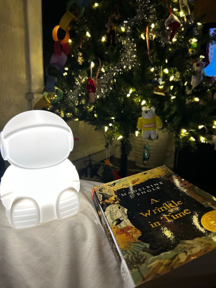 An astronaut nightlight and the book “A Wrinkle in Time” sit on the corner of the bed with a lit Christmas tree in the background. 