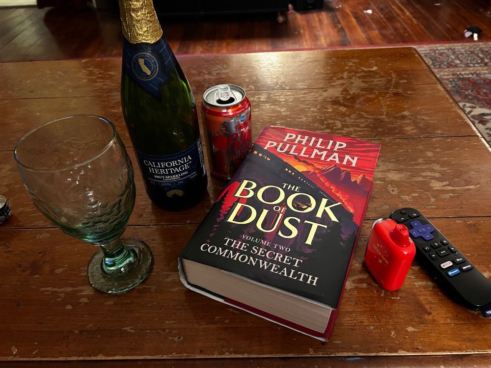A bottle of brut, wine glass, sparkling water, vape, Roku remote and The Book of Dust by Philip Pullman on the coffee table