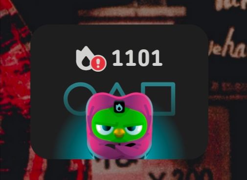 Screen cap of the Duolingo widget on Huyen's desktop. The Duolingo owl is dressed a guard from the television show "Squid Games" with the streak fire emoji as its emblem while the familiar Squid game Circle, Triangle, and Square sit behind the owl, lit in blue with a spotlight seemingly shining on the viewer as they attempt to escape the cruel game of maintaining Diamond League every week.