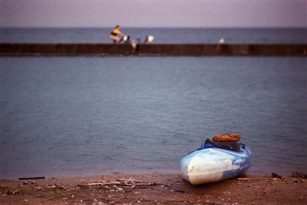 A white and blue kayak is pulled up on the beach. In the distance, out of focus and beyond the breakwater, two people ride by on a pedalboat. 