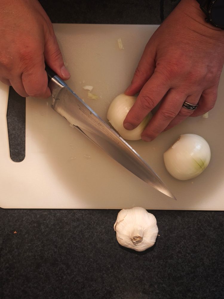 Das Bild zeigt eine Person, die auf einem weißen Schneidebrett Zwiebeln schneidet. Die Person benutzt ein großes, scharfes Messer, um die Zwiebeln zu schneiden. Auf dem Brett liegen bereits einige geschnittene Zwiebelstücke. Unterhalb des Schneidebretts liegt eine Knoblauchknolle auf einer dunklen Arbeitsfläche. Die Person trägt einen Ring am Finger und eine Smartwatch am Handgelenk (KI Text) 