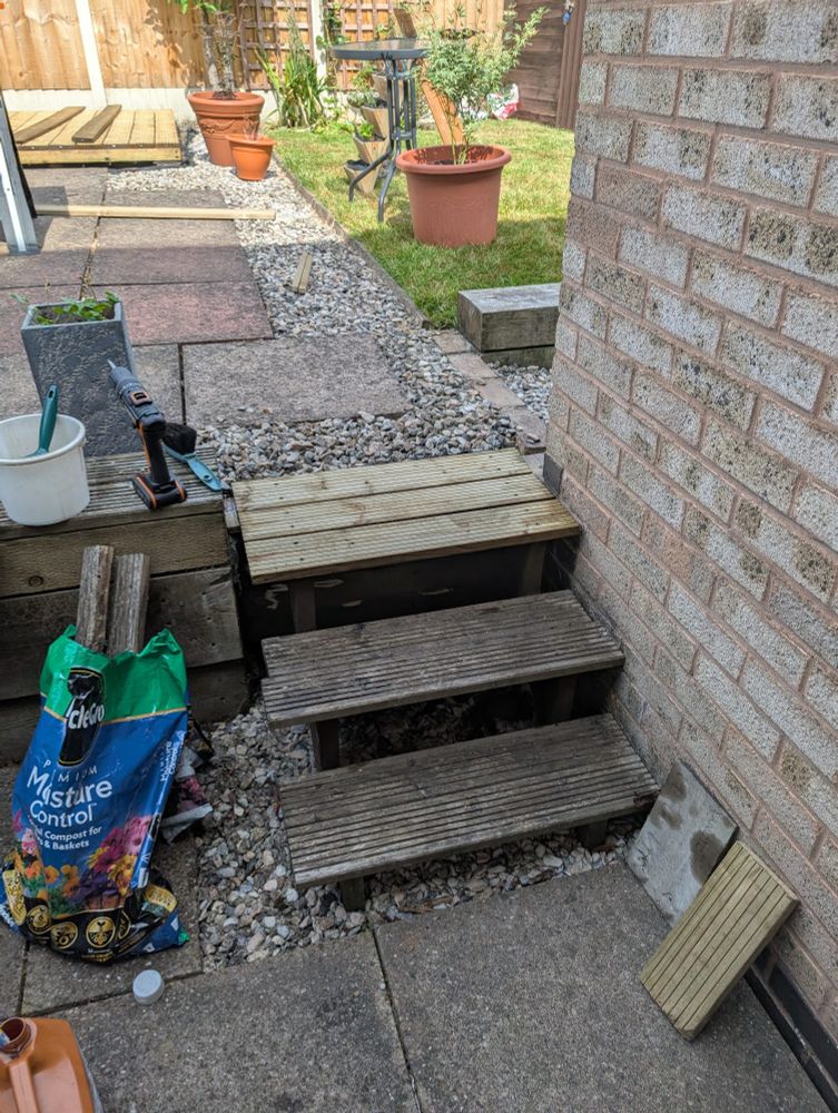 A photo of a wooden stairs making up 3 steps, going from a lower part of the garden to the upper part. The top steps have been replaced with new looking decking planks. The stairs lead from a patio to an upper patio