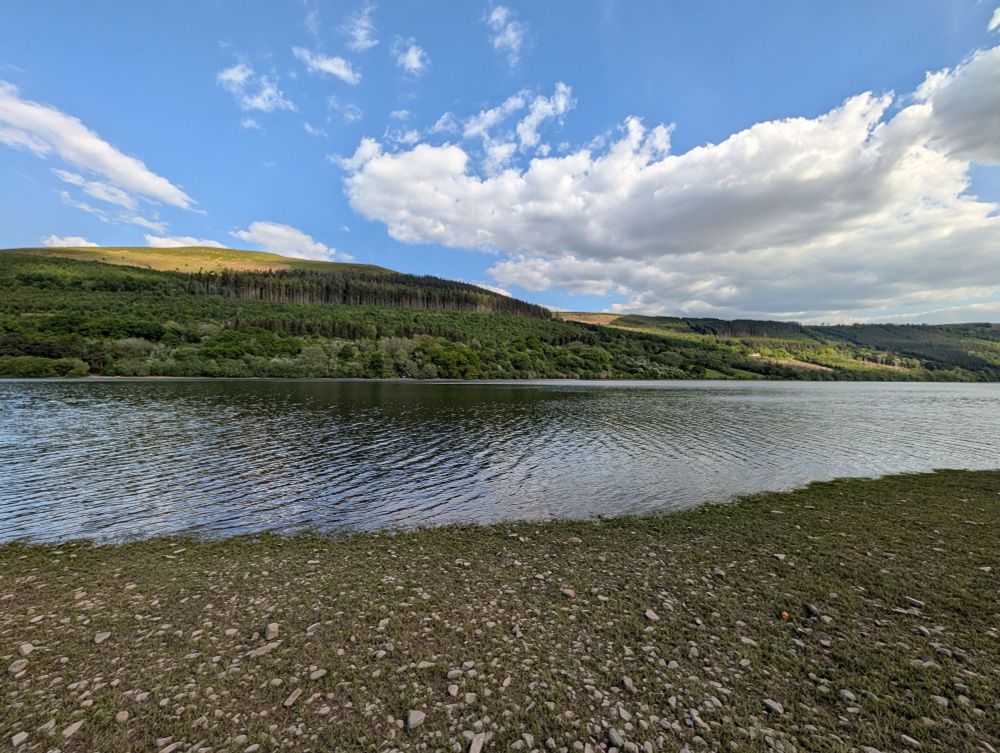 Photo of a reservoir with blue skies and light clouds. There is a gravel beach and rolling hills with forests on them