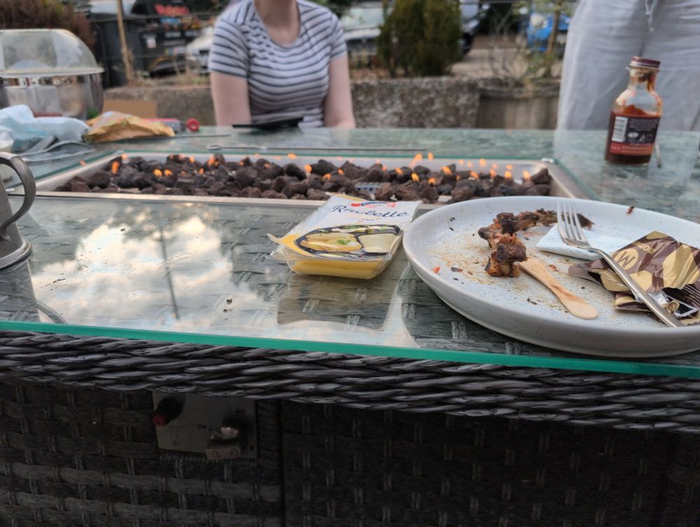 Picture of a glass table with a fire in the middle. There in a plate on the table with eaten food on it, and a person in a stripy shirt sitting behind the table