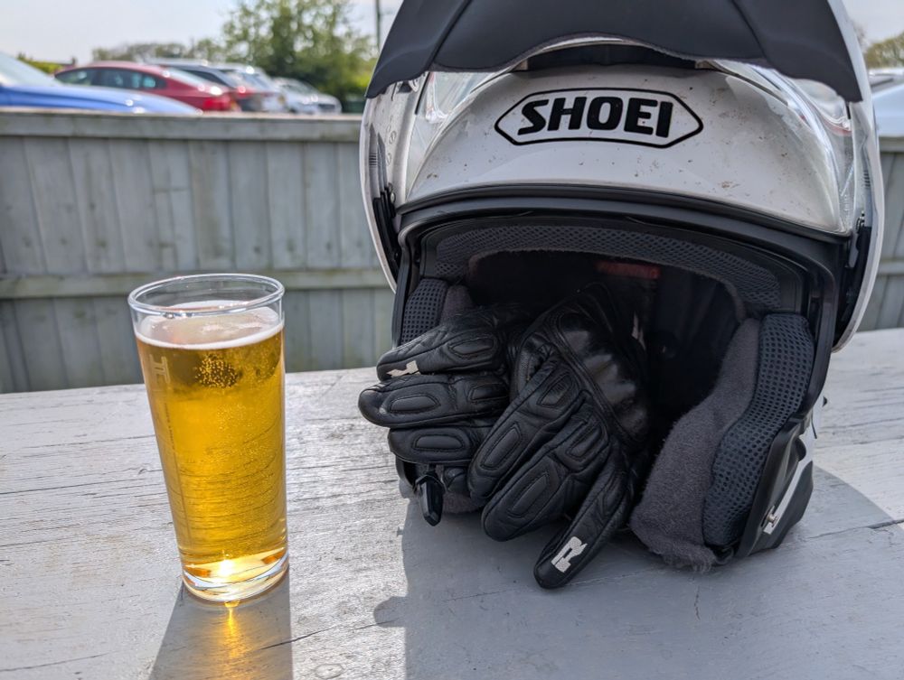 A photo of a half pint of cider next to a white motorbike helmet with a pair of black gloves inside the helmet. They are sitting on top of a grey picnic table, in a sunny setting