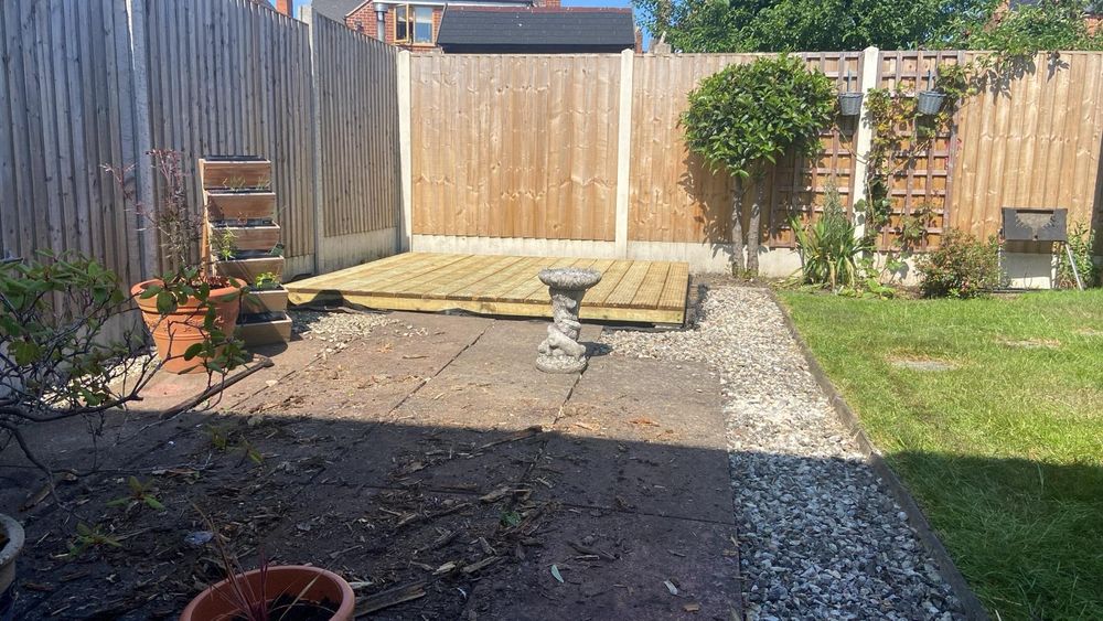 An outside photo of a garden. There is a patio at the front, with plant pots to the left and a grass patch to the right. At the back of the garden is a newly laid decking