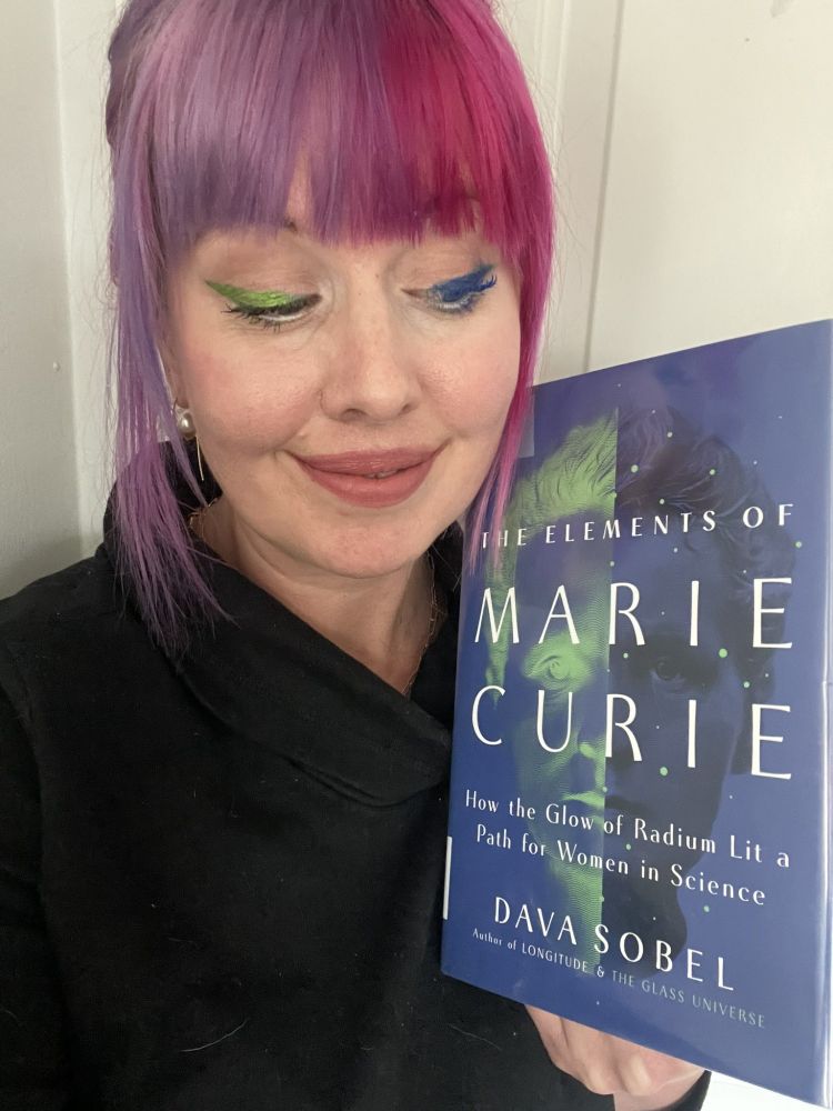 Image description: A woman with pink and purple hair with green and blue eyeliner holds a copy of the book THE ELEMENTS OF MARIE CURIE: HOW THE GLOW OF RADIUM LIT A PATH FOR WOMEN IN SCIENCE by Dava Sobel. 

