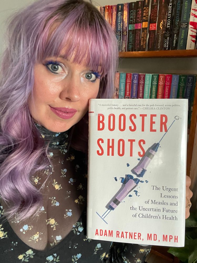 Selfie of a white woman with pink and purple hair holding the book BOOSTER SHOTS by Adam Ratner MD