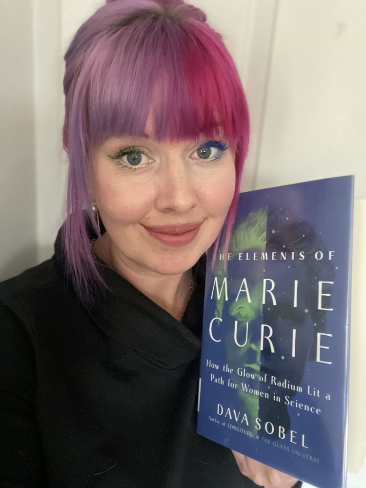 Image description: A woman with pink and purple hair with green and blue eyeliner holds a copy of the book THE ELEMENTS OF MARIE CURIE: HOW THE GLOW OF RADIUM LIT A PATH FOR WOMEN IN SCIENCE by Dava Sobel. 
