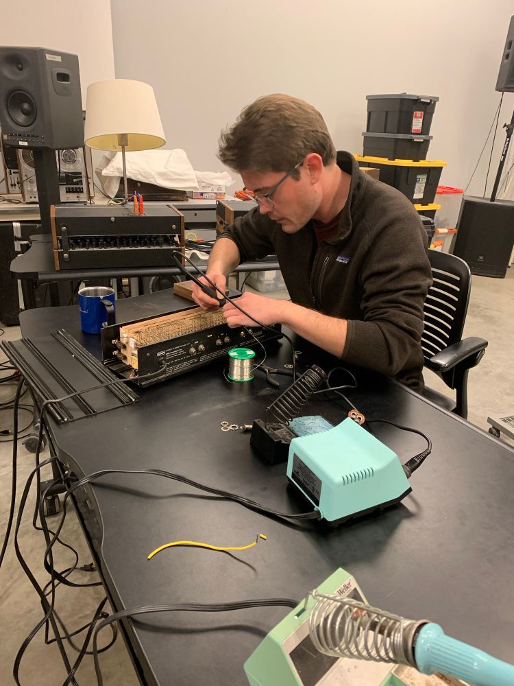 A young man wearing glasses, appearing slightly stressed out, leans over a table holding a solder iron. He is poking around in the guts of some sort of audio equipment. 