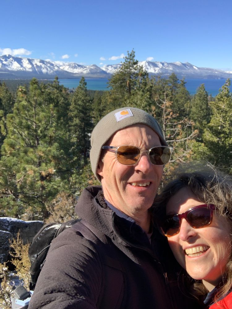 A middle-aged man and woman wearing sunglasses squint at the camera while taking a selfie. They're outdoors, with pine trees in the background, and in the far distance, a big lake and snow-capped mountains. The man is wearing a warm hat. They're on a hike in the woods. 