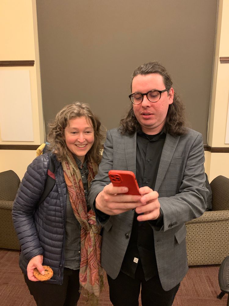 Two people in a green room backstage get ready to take a picture. A man is holding his phone and trying to figure out how to flip the camera, while a woman wearing a scarf stands next to him and grins. He looks a little stressed out but she is having a good time. 