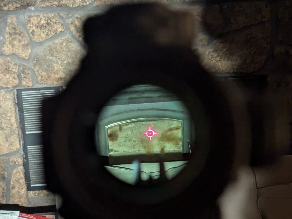 Picture of an indoor fireplace, taken thru the sight of a rifle. A red reticle is visible over the fireplace door.