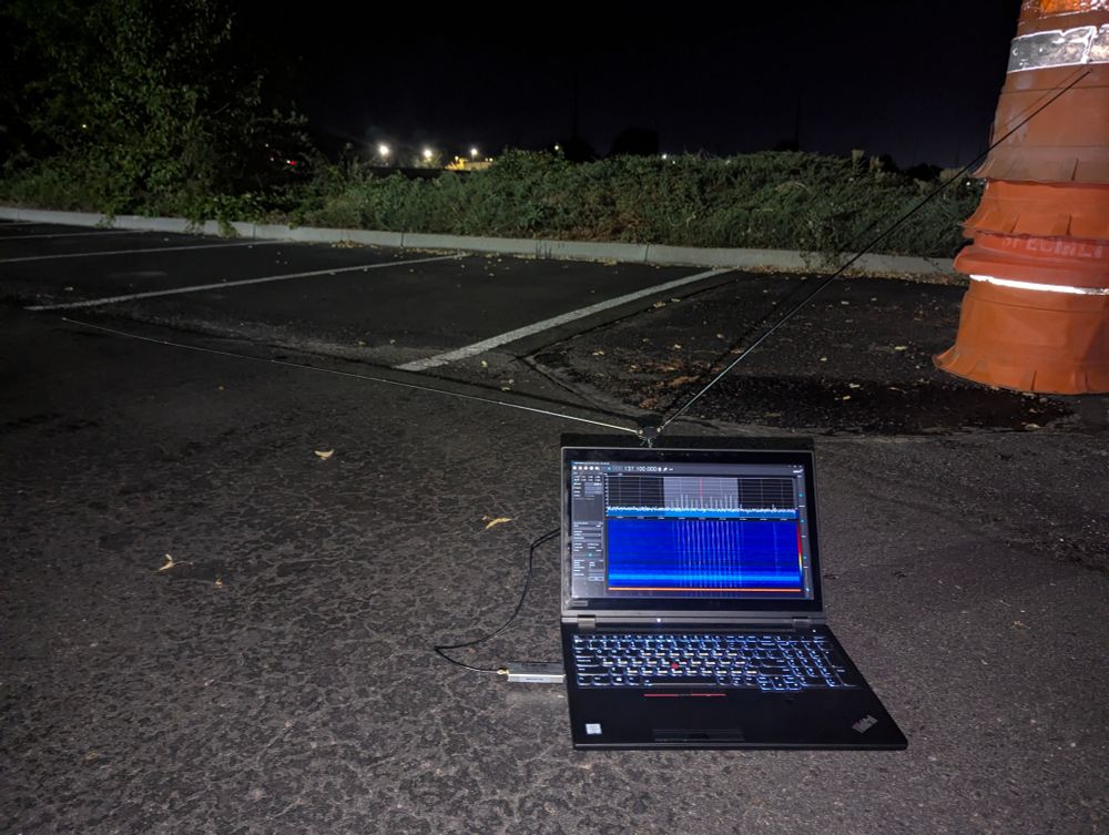 Picture of a ThinkPad laptop sitting in a dark parking lot, with a 2m antenna attached to the back. The laptop is running SDR#, tuned to 137.1MHz