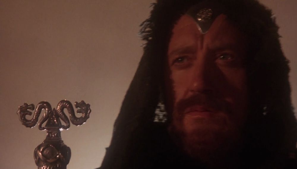 Nicole Williamson as Merlin holds a staff with a two-headed dragon on the end. 