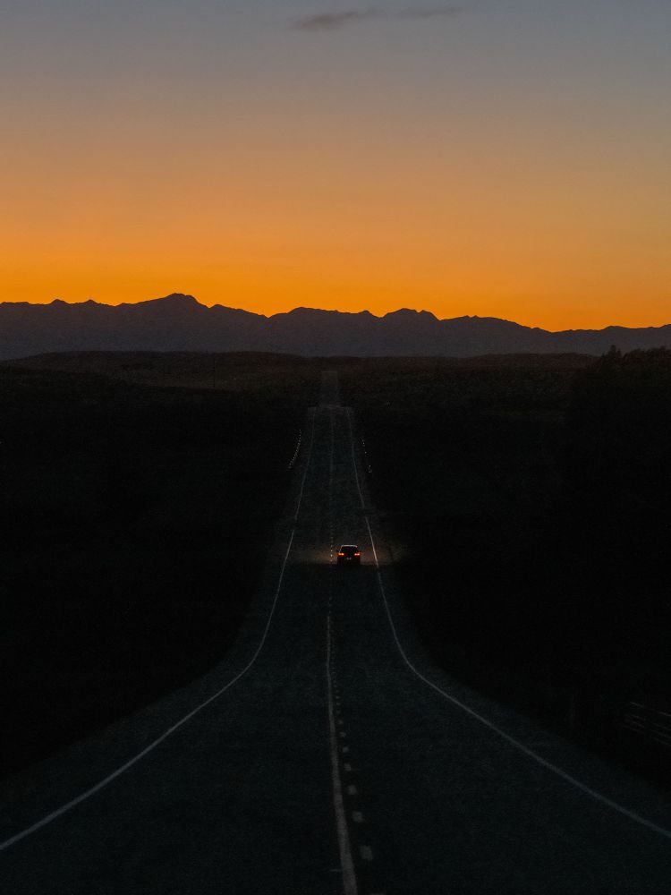 Post blue hour near mountains. A car drives away on two lane roadway, illuminating it. 