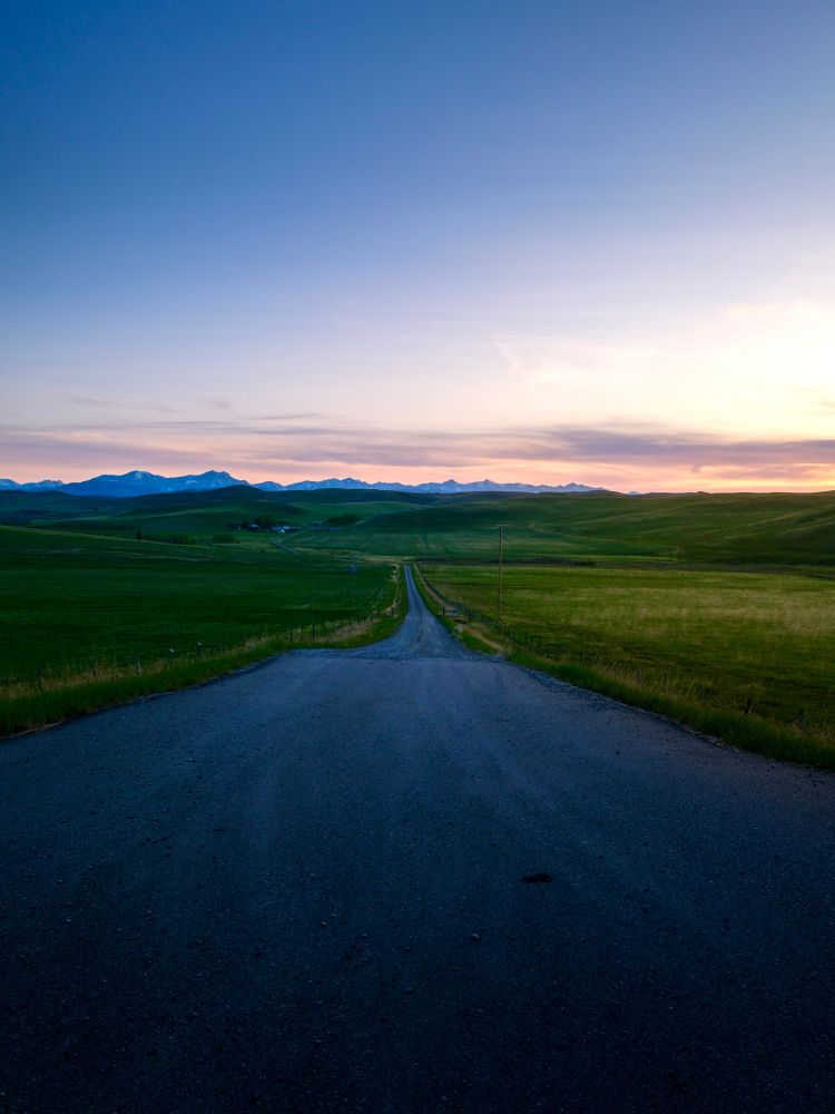 A dirt road across a green field into a valley. Rocky Mountains of Kananaskis in the distance. GFX100RF
