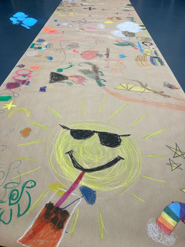 Eine Bahn Packpapier, die von Kindern bemalt wurde. Vorne im Zentrum befindet sich eine Sonne mit Sonnenbrille (vor was schützt sie sie eigentlich…?), die über einen Trinkhalm etwas Rötliches aus einem Glas trinkt. 