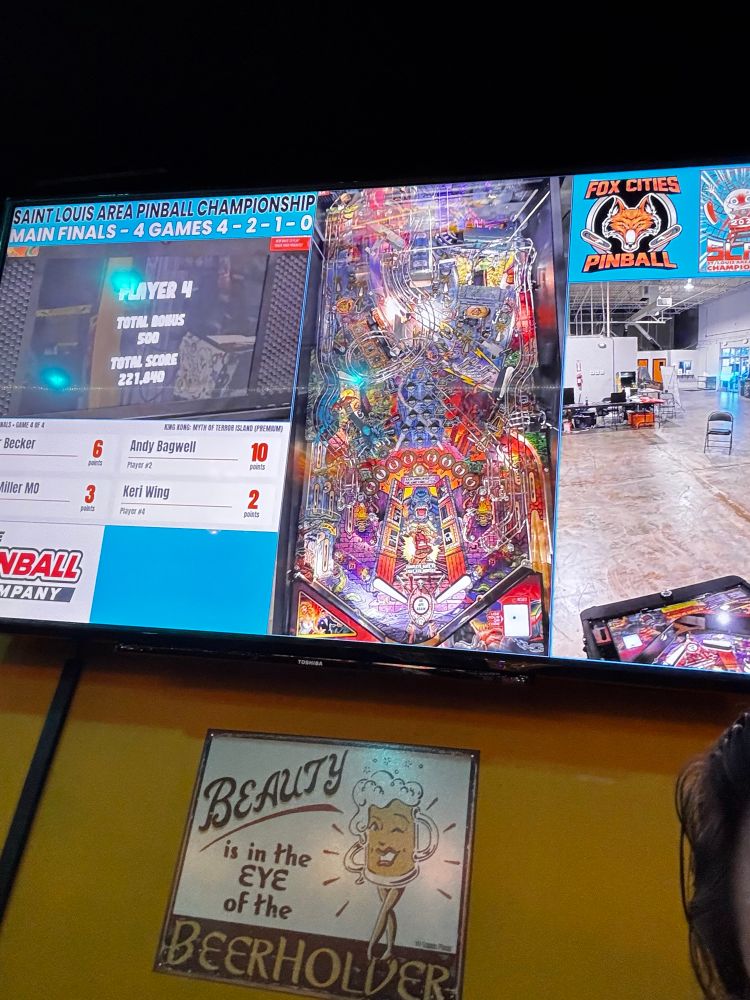 Tv showing live play from the St. Louis Area Pinball Championship 
It shows an overhead of the King Kong pinball game next to a scoreboard and a dashcam view of the players

Beneath the tv is a sign on the wall that says “beauty is in the eye of the beerholder”