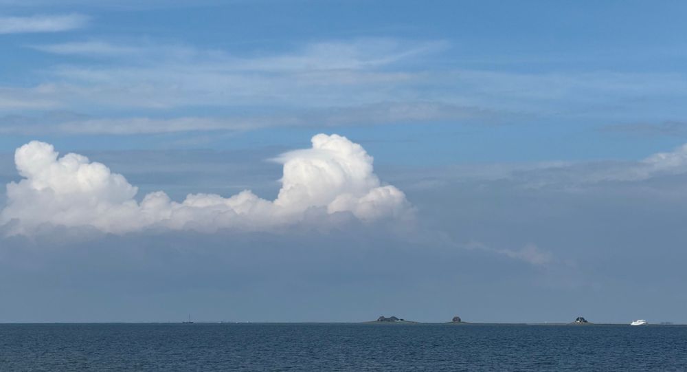 The North Sea under some clouds. On the horizon the buildings on Hallig Nordstrandischmoor are visible. A white Ferry is on the right side.