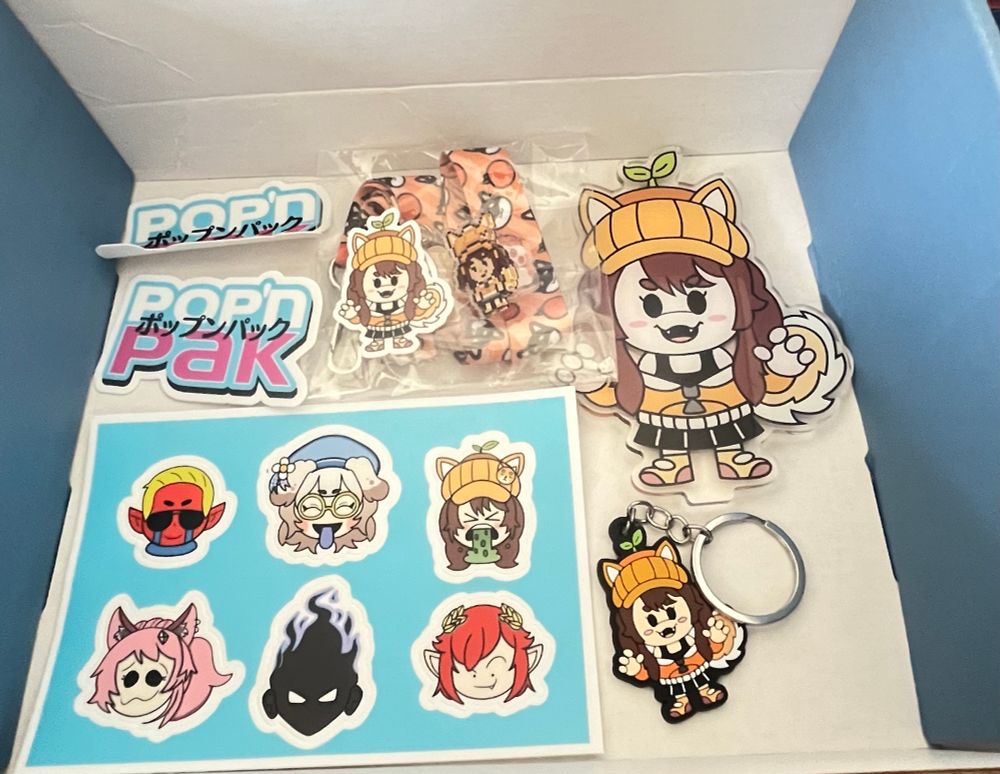 A Pop'nPak Box filled with Aki (OrangeisBorange) Merch. There's 2 Pop'nPak Stickers, an OrangeisBorange Acrylic Keychain + Lanyard, Small Die Cut Sticker, Acrylic Pixel Pin, Rubber Keychain, Acrylic Standee & 6 Stickers of Aki & her friends.