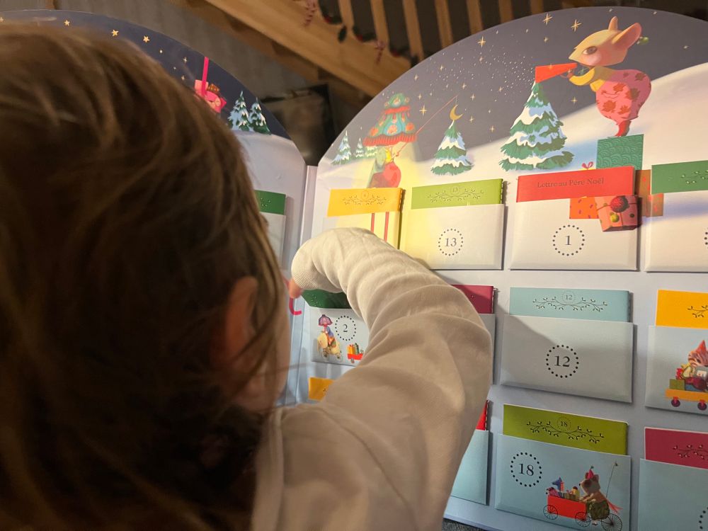 Photo de dos d’un enfant qui a des cheveux châtain clair Lenfant met sa main dans la case 2 du calendrier de l’avent (ce sont de petites cases dans lesquelles se trouvent des mini livres)