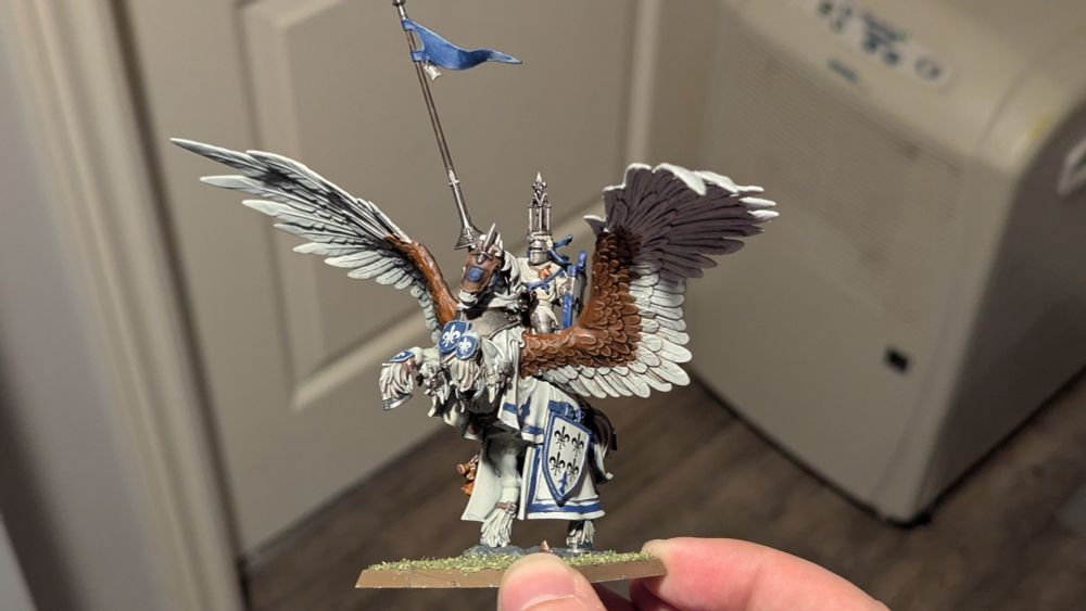 Bretonnian Lord on Royal Pegasus. White horse with white and brown eagle wings rearing up with a white clad knight on its back. The ride holds a lance aloft with a blue pennant at the end. The Pegasus is adorned with Bretonnian fleur-de-lis heraldry. Photographed from the right side.