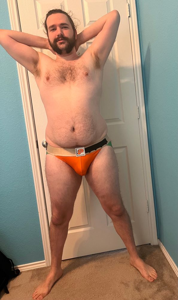 Full body of Austen in an orange jockstrap with green straps, with their hands behind their head