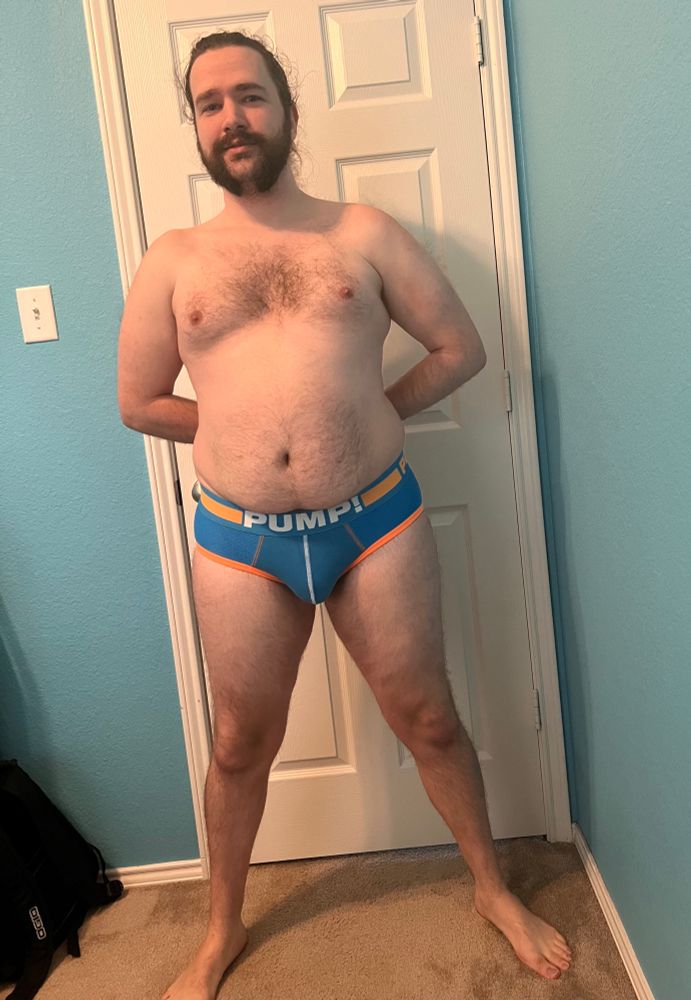 Full body of Austen facing forward in blue Pump briefs with orange accents, arms behind their back