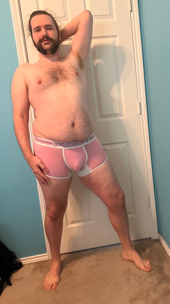Full body front view of Austen in pink Pump trunks with white accents, right arm at their side and left hand behind their head
