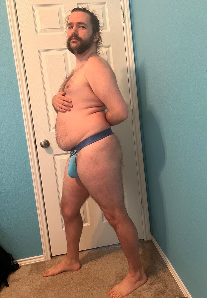 Full body left-side view of Austen in a light blue jockstrap with a blue waistband, right hand above their stomach and left arm behind their back