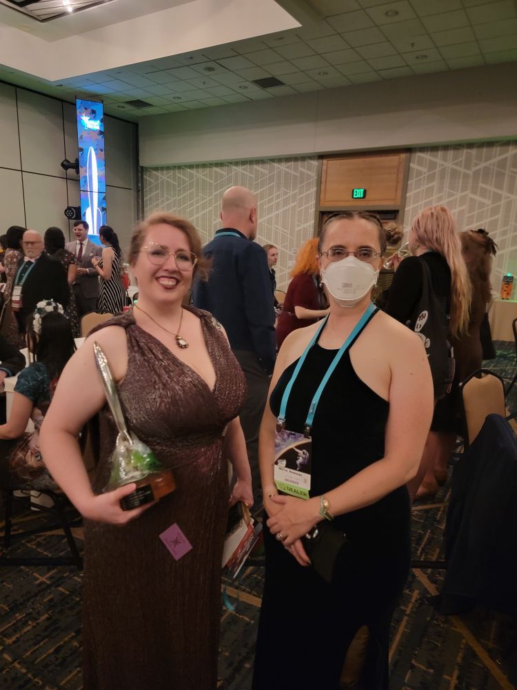 Animator and director Megan C. Lloyd and author and poet Marie Brennan in formal dresses at the 2025 Hugo Awards after party.  Megan is holding a Hugo in her right arm.