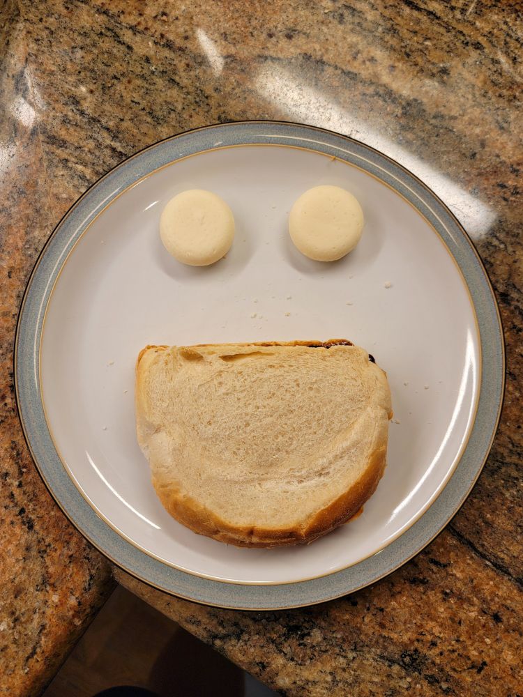A plate is centered in the photo, arranged with a sandwich and two unwrapped Babybel cheeses. The sandwich is at the bottom of the plate, oriented with the flat part of the bread crust toward the top of the plate.  The Babybels are toward the top of the plate, side-by-side.  The overall arrangement is suggestive of a smiling face.