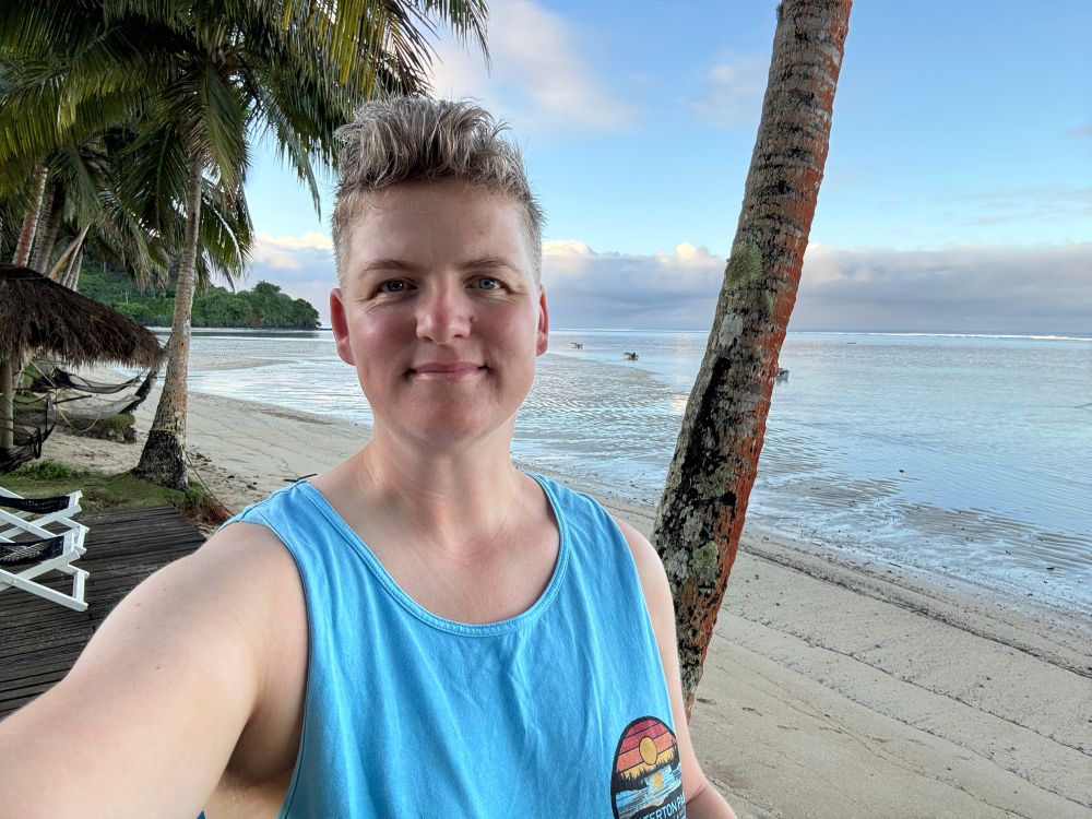 Selfie av person på strand i en blå singlet med palmetrær i bakgrunnen. 