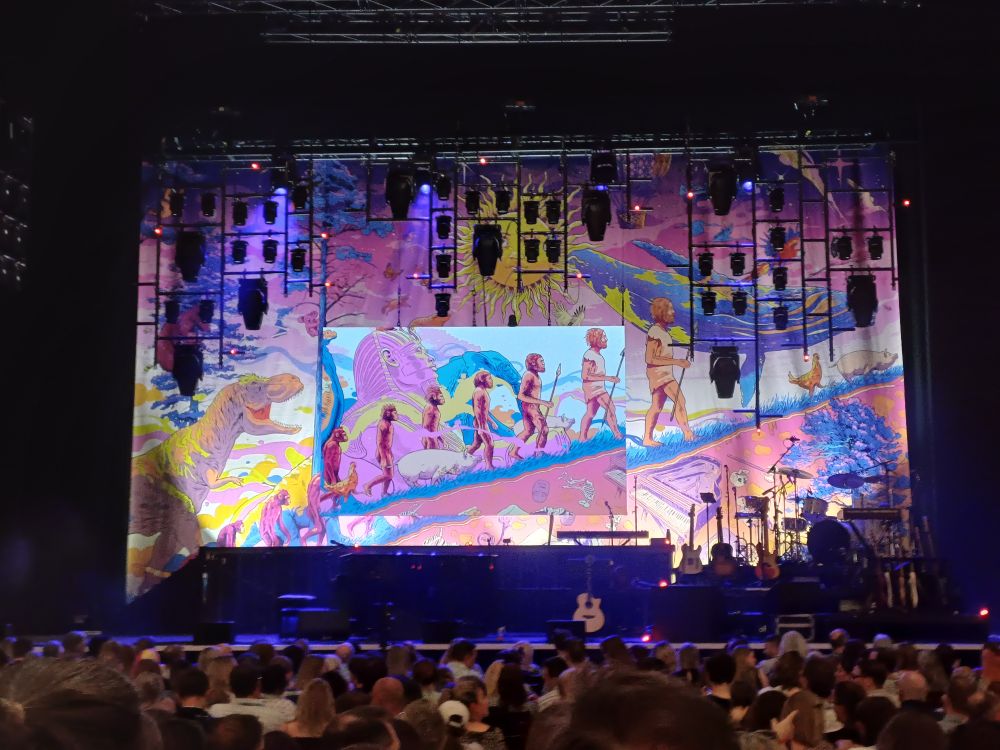 The empty stage at Sheffield City Hall. There are a large number of instruments on stage including about 10 guitars, a drum kit and a baby brand piano. The backdrop is a cloth screen artwork with the evolution of humans in the centre, a t rex to the left and various animals and instruments scattered around
