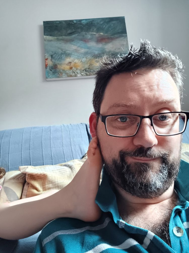 Selfie of Rob sitting on the ground with a blue-grey couch in the background and an abstract painting on the wall. There is a small child's foot resting on his shoulder, resting against his cheek