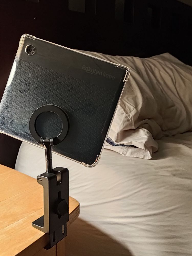View from the side of a bed of an ereader mounted on a magnetic stand that is attached to the nightstand table. The ereader is angled for optimal viewing for someone laying down on the pillow on the bed just beyond the ereader 