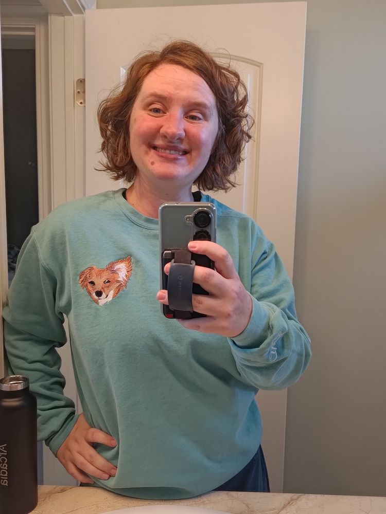 A 30 year old woman with chin-length wavy red hair taking a mirror selfie wearing a teal crew sweatshirt depicting an embroidered image over the woman's heart of a little red dog with one ear sticking up.