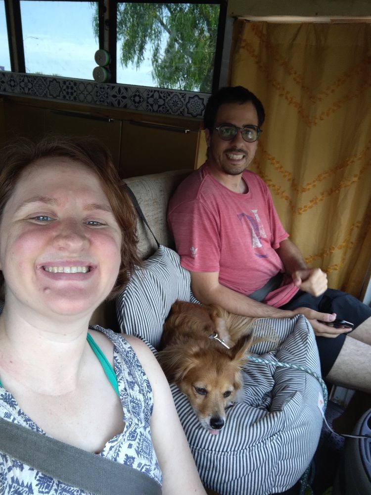 A woman, a man, and a small mixed breed dog smiling at the camera, sitting in seats with seatbelts on inside a motorhome with a gold curtain and cabinets behind them. The dog has a delightful little blep.