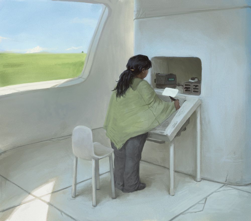 A woman in a poncho stands in a plain room at a drawing table holding some kind of tool. On a shelf are some tools and an old radio