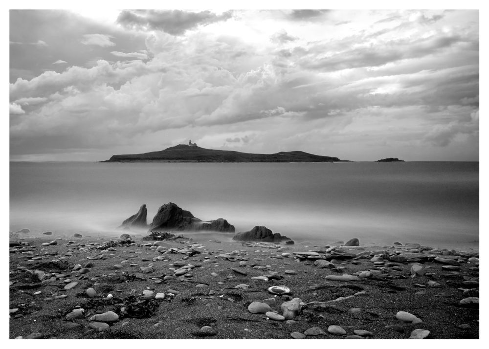 A serene coastal landscape with a rocky foreground and a pebbled beach leading to calm, misty waters. An island with a lighthouse is visible in the distance under a cloudy sky.