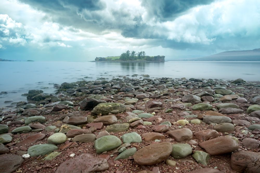 A rocky shoreline with various-sized stones leads to a calm body of water. In the distance, a small green island lies under a cloudy, overcast sky. The landscape reflects a tranquil atmosphere.