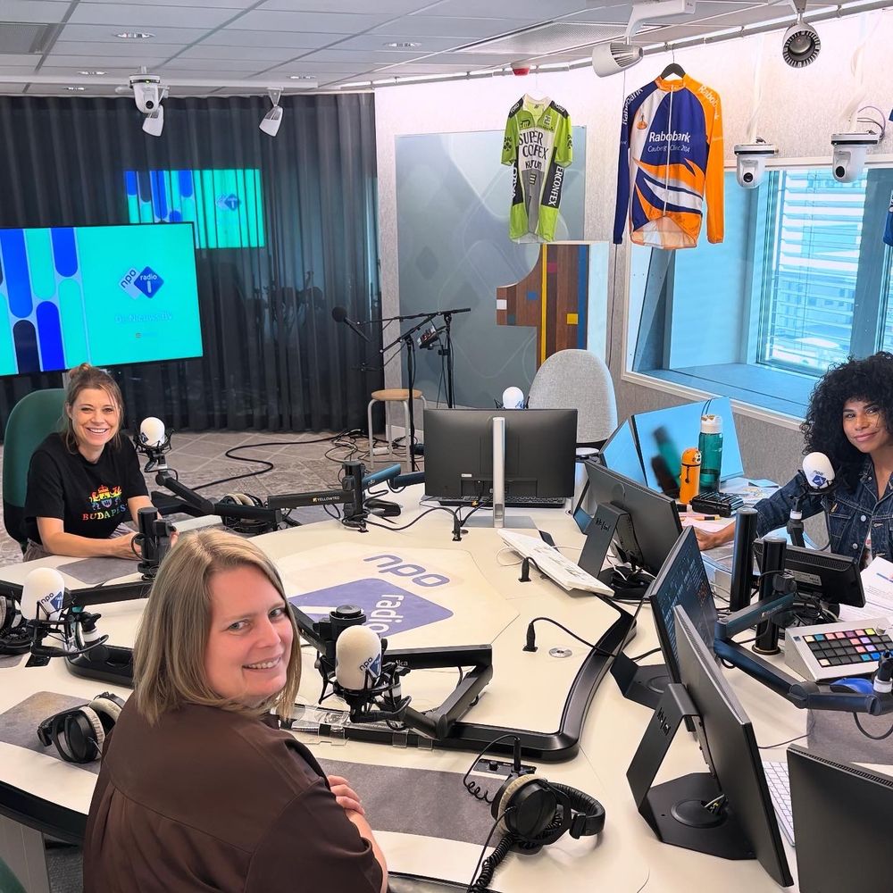 Foto van de studio van De Nieuws BV waar ik (linksachter de tafel) met Berdien (vooraan op de foto), Natasja Gibbs (rechts) achter een driehoekige witte tafel zit met in het blauw het NPO1 radio logo in het midden en allerlei microfoons, koptelefoons en snoeten zit.