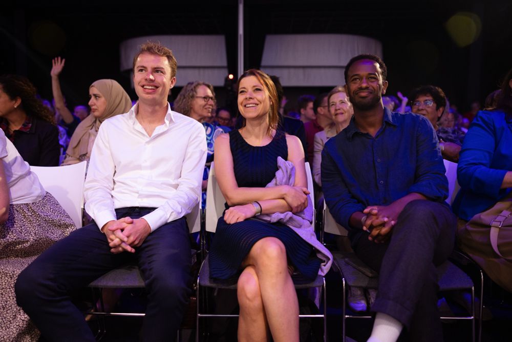 Foto van v.l.n.r. Julian (witte blouse, donkere broek), ik (blauwe jurk) en Habtamu (donkerblauwe blouse en zwarte broek). We zitten op de voorste rij van het congres, daarachter veel mensen. 